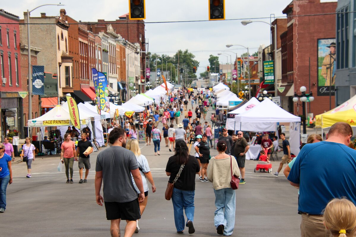 Street festival with people walking among vendor tents on a downtown street lined with historic brick buildings.