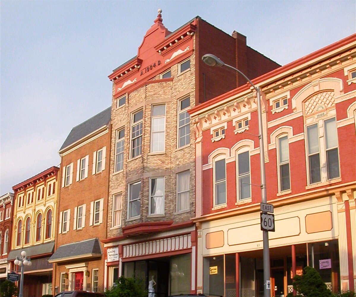 A row of historic brick buildings in a small downtown area, showcasing 19th-century architectural styles. The buildings have ornate cornices, tall windows, and painted facades in shades of red, tan, and cream. One building has “A.D. 1904” inscribed near its roofline. A lamppost and a U.S. Route 60 highway sign are visible along the sidewalk in front of the storefronts.