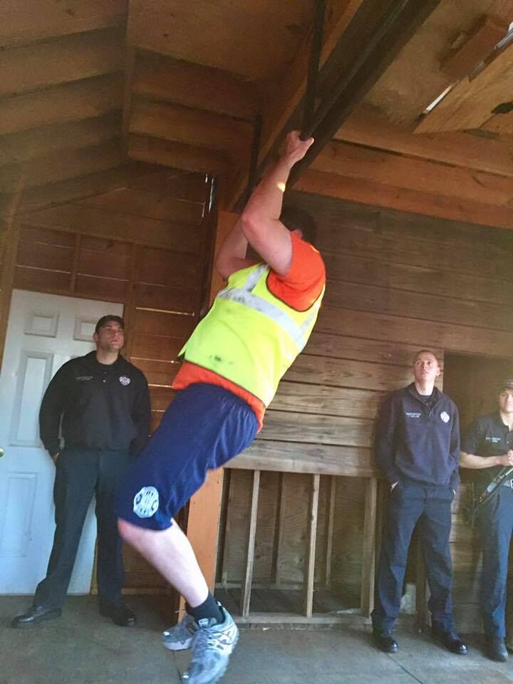 Person wearing an orange shirt and reflective vest performing a pull-up on a metal bar inside a wooden structure. Several individuals in dark uniforms stand in the background near a white door and unfinished wooden walls.