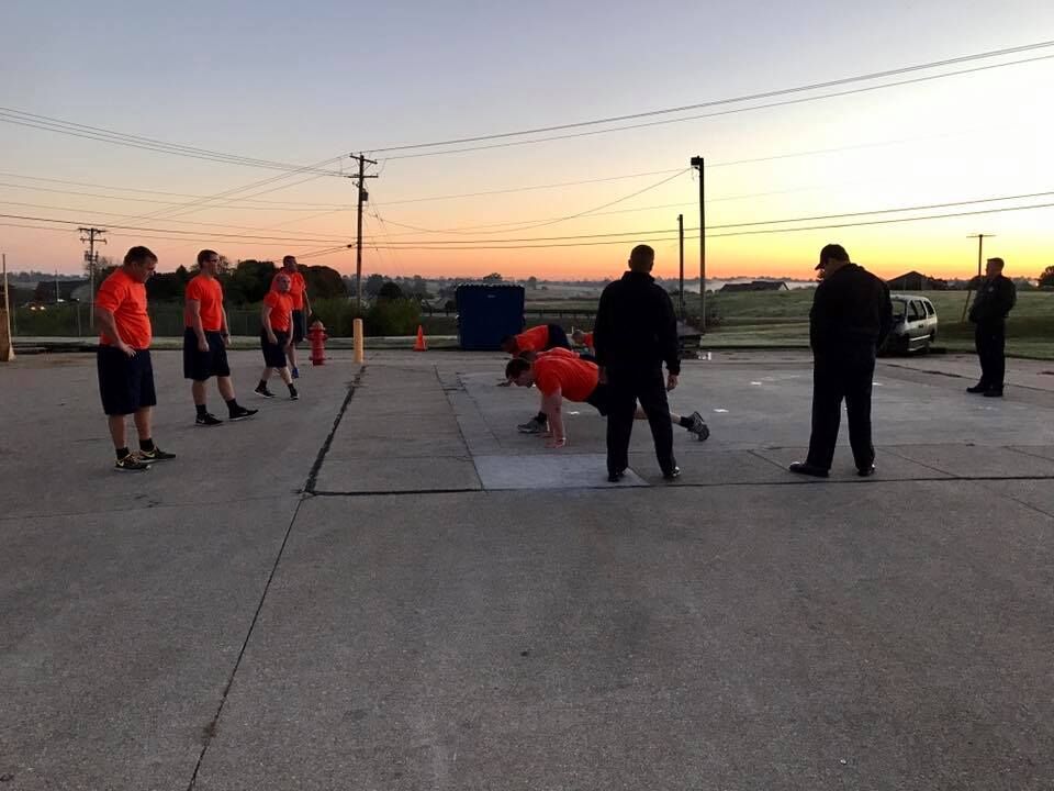 Group of individuals wearing matching orange shirts performing outdoor physical fitness drills on a paved surface at sunrise. Some participants are standing while others are in a push-up position. Two people in dark uniforms observe nearby. Utility poles and a vehicle are visible in the background.