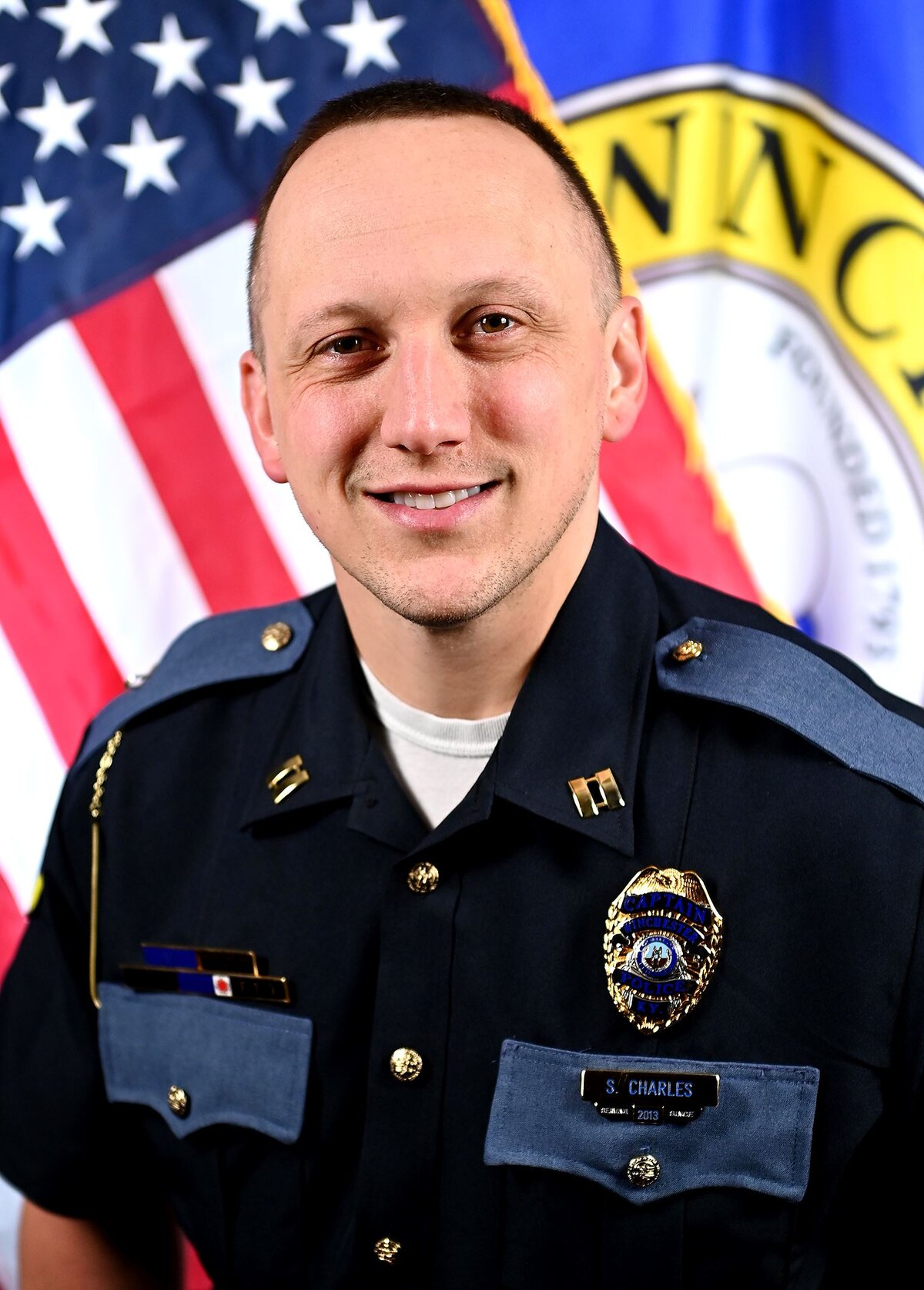 Person wearing a dark formal uniform with shoulder epaulets and a badge that reads “S. Charles.” The uniform includes gold pins and a nameplate. In the background, there is an American flag and part of a circular emblem with visible text “Winchester” and “Founded 1793.”