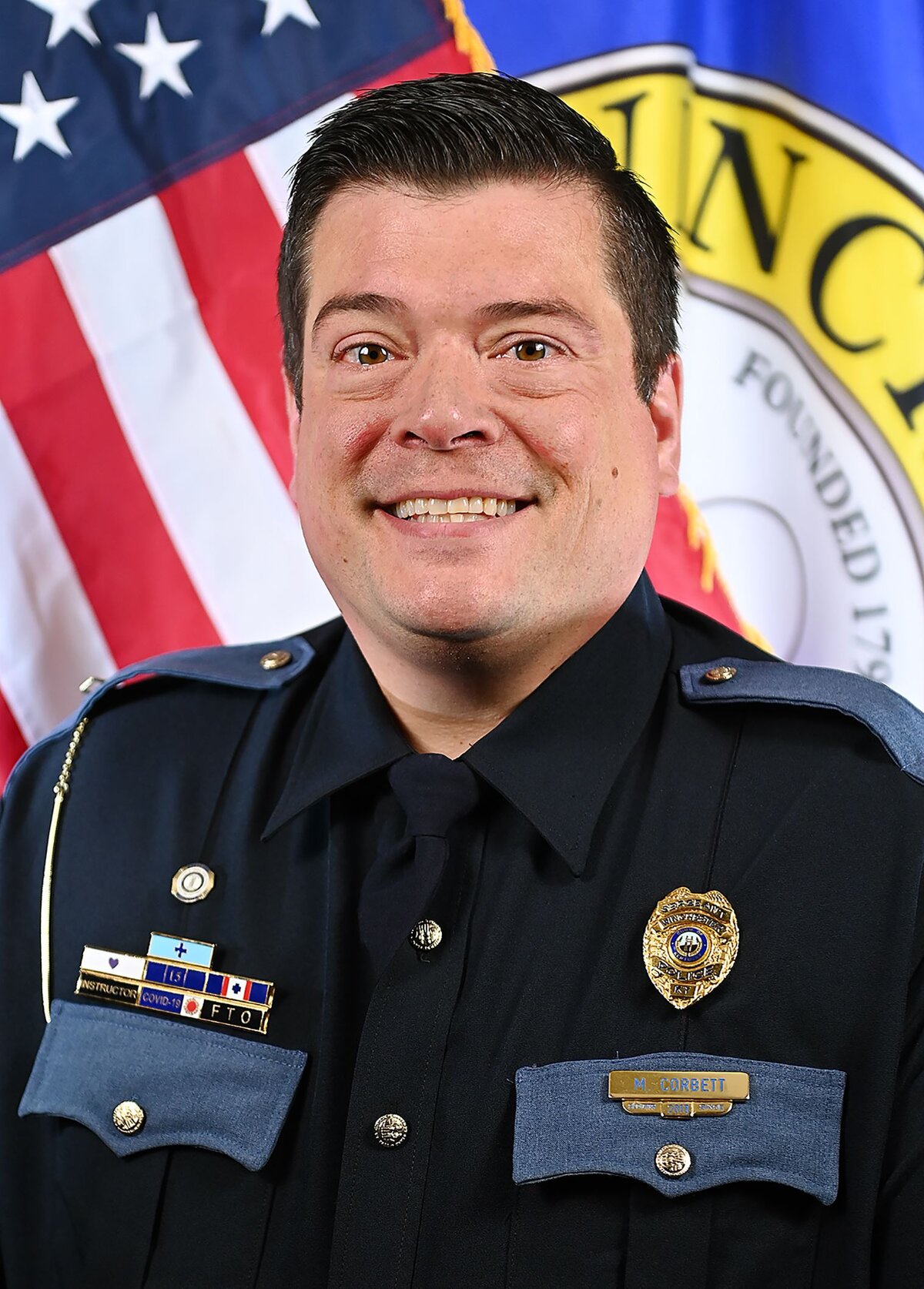 Person wearing a dark formal uniform with shoulder epaulets, a tie, and a badge. The uniform includes a nameplate reading “J. Garrett” and several service ribbons. In the background, there is an American flag and part of a circular emblem with visible text “Winchester” and “Founded 1793.”
