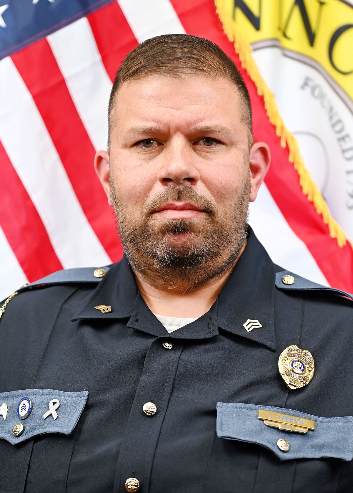 Person wearing a dark formal uniform with gray shoulder epaulets and silver buttons. The uniform includes a badge and a nameplate reading “J. Thomas,” along with service pins and ribbons. An American flag and part of a circular emblem with visible text “Winchester” and “Founded 1793” are in the background.