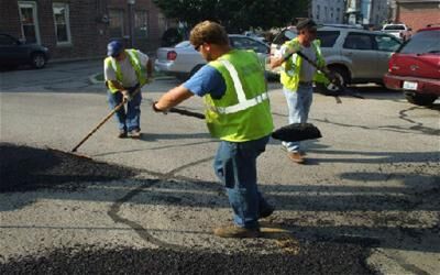 Three workers wearing reflective safety vests spreading asphalt on a street using shovels and rakes. Vehicles and buildings are visible in the background.