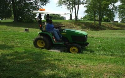 Person operating a green John Deere tractor with a canopy on a grassy slope in a park-like setting with trees in the background.