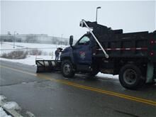 Blue dump truck equipped with a snowplow clearing a snow-covered road. Snow is visible on the ground and in the surrounding area.