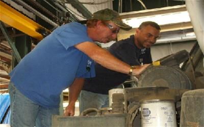 Two individuals inspecting and working on machinery inside a maintenance facility. Various mechanical parts and tools are visible.