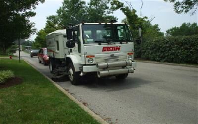 White street sweeper truck parked on a paved road with a red vehicle behind it. Trees and bushes line the street.
