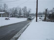 Snow-covered residential street with a vehicle driving toward the camera. Trees, utility poles, and houses line the road.
