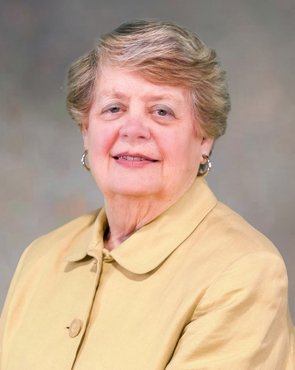 Portrait of an individual wearing a light tan jacket with a rounded collar and button detail. The person has short, neatly styled hair and is wearing hoop earrings. The background is a neutral gradient.